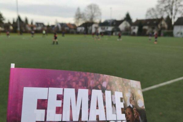 Nahaufnahme eines Flyers mit der Aufschrift &bdquo;FEMALE&ldquo; im Vordergrund, im Hintergrund spielt eine Frauenfu&szlig;ballmannschaft.