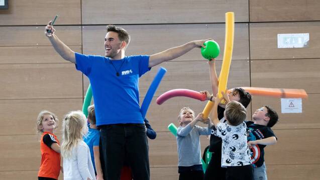 Ein Trainer mit einem Ball steht umgeben von Kindern, die bunte Schwimmnudeln in einer Sporthalle halten.
