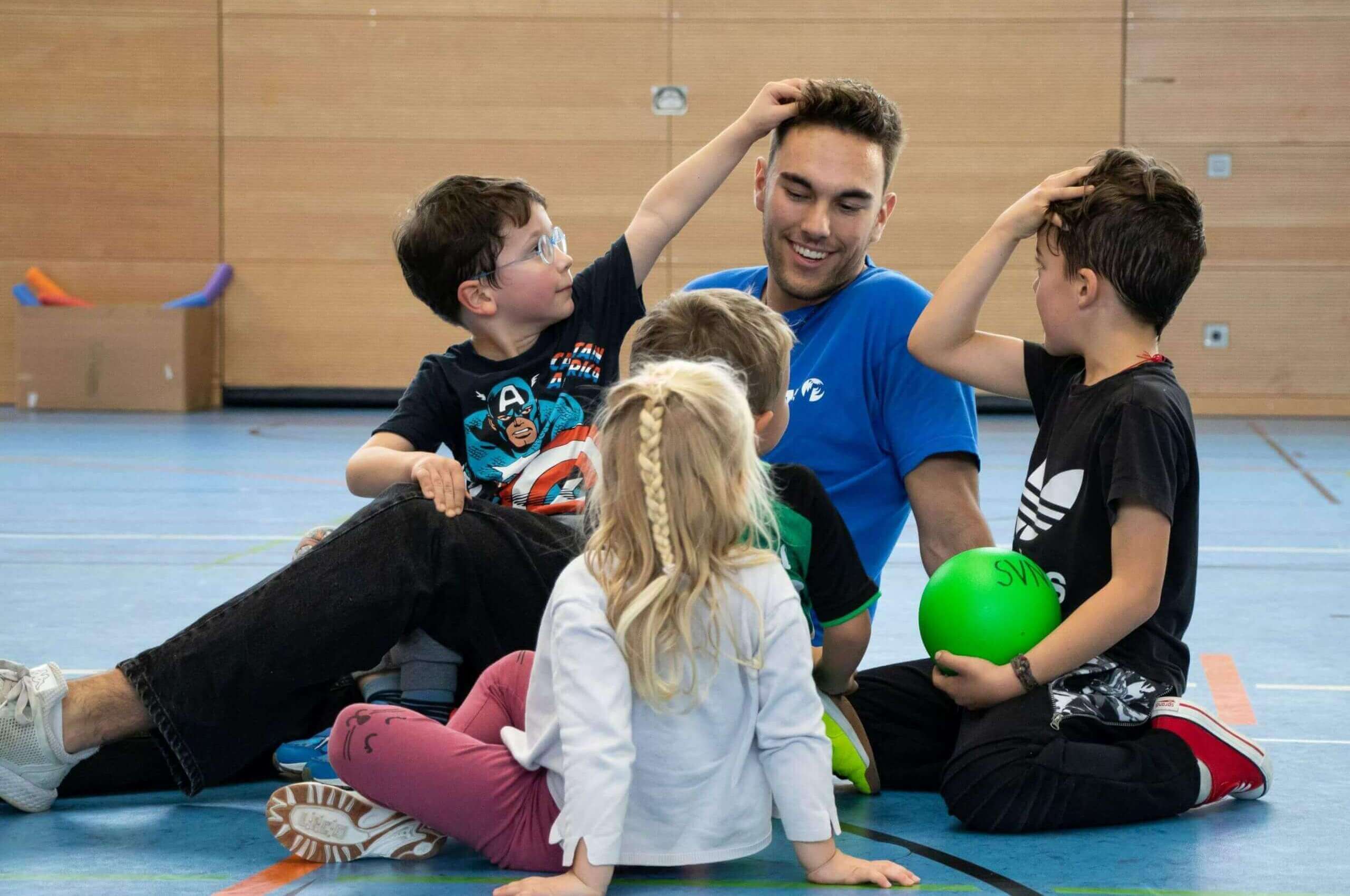 Ein junger Mann sitzt mit vier Kindern auf dem Boden einer Sporthalle, w&auml;hrend sie miteinander spielen und lachen.