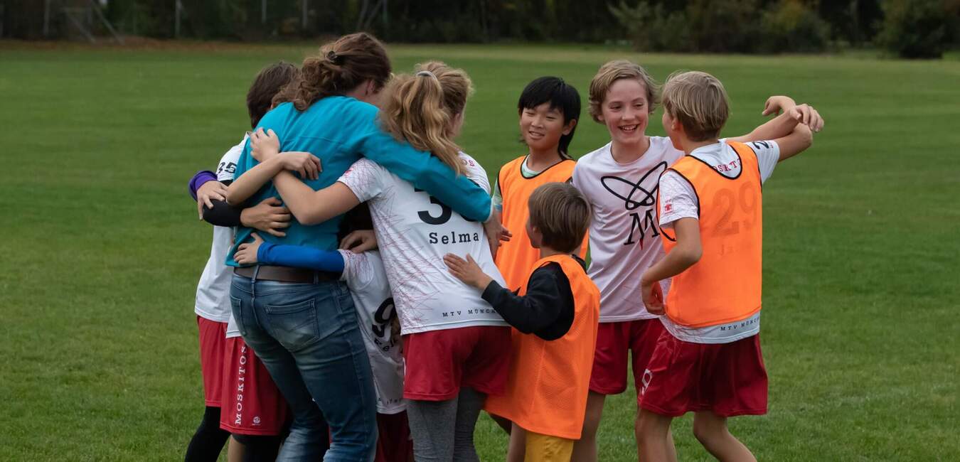 Gruppe von Kindern in Fußballtrikots umarmt sich freudig auf einem grünen Sportfeld.