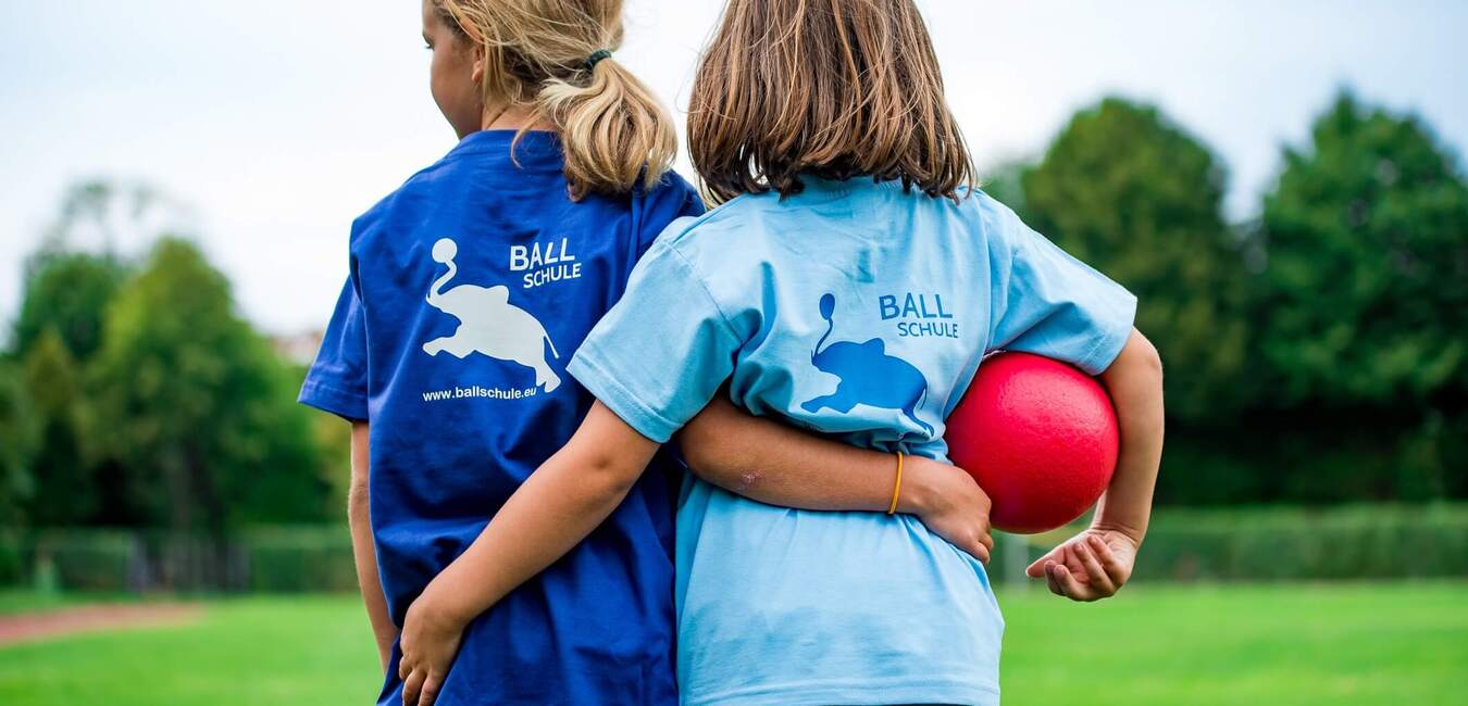 Zwei Kinder stehen Rücken an Rücken auf einem grünen Feld, eines hält einen roten Ball und trägt ein blaues T-Shirt.