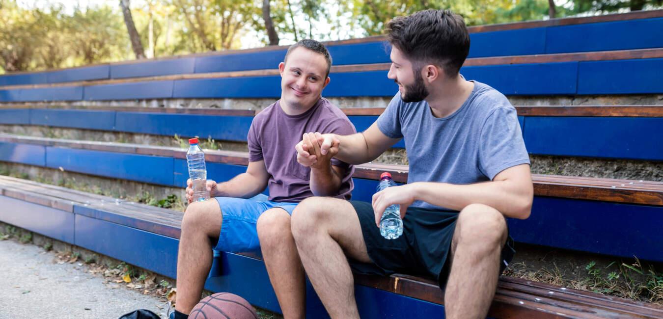 Zwei M&auml;nner sitzen auf einer Trib&uuml;ne, einer zeigt auf den anderen; Basketball und Wasserflaschen liegen neben ihnen.