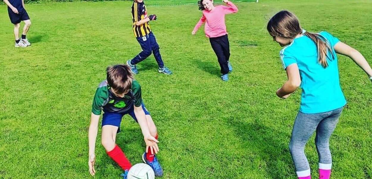Vier Kinder spielen auf einem Fußballfeld, während einer den Ball dribbelt und die anderen ihn verfolgen.