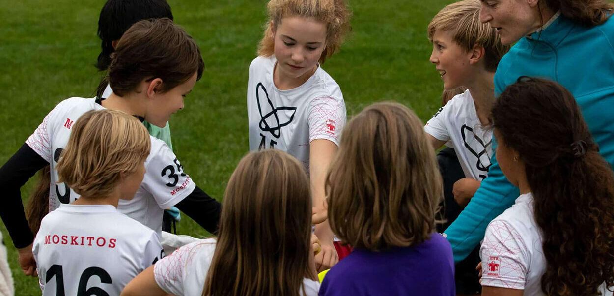 Gruppe von Kindern in Sporttrikots um einen Ball, w&auml;hrend sie sich vor einem Spiel motivieren.