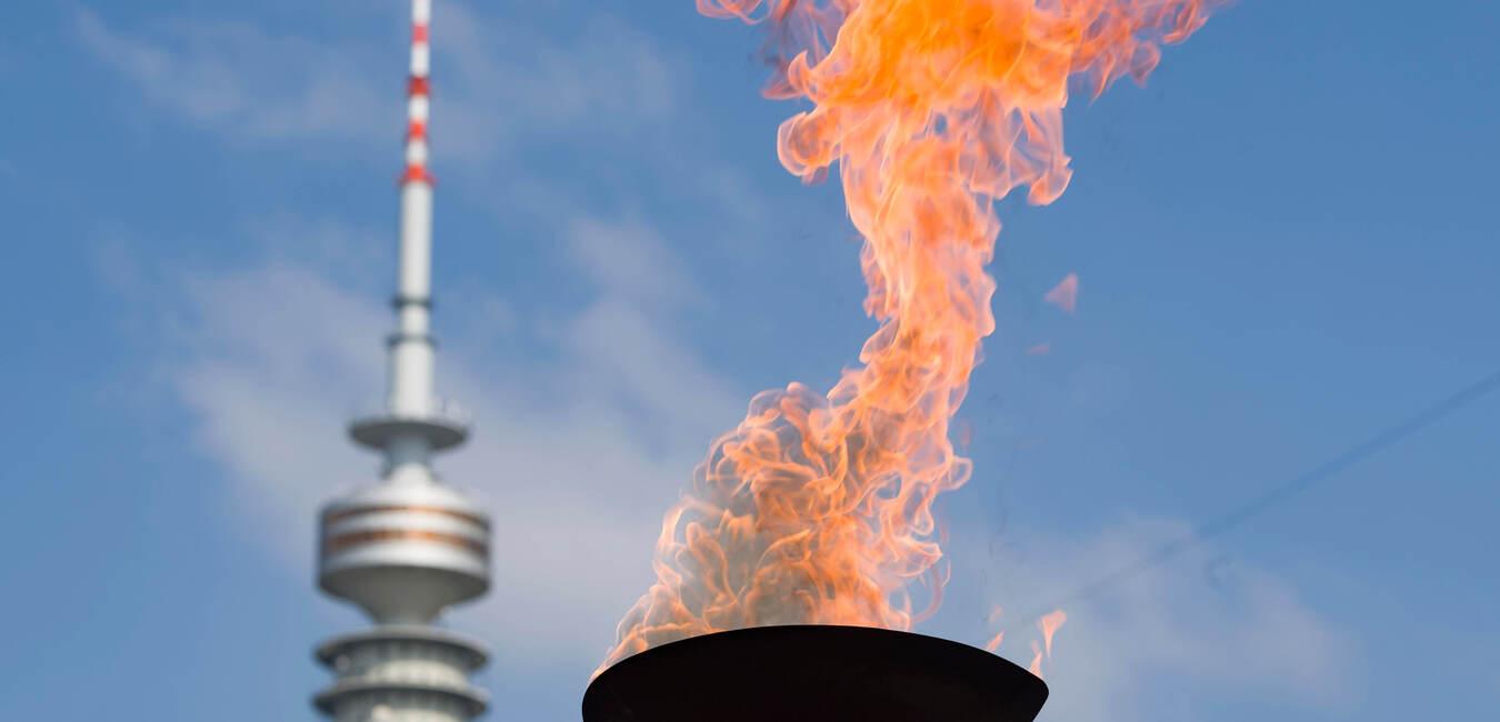 Flamme in einer Schale vor einem Fernsehturm, unter blue Himmel mit wenigen Wolken.