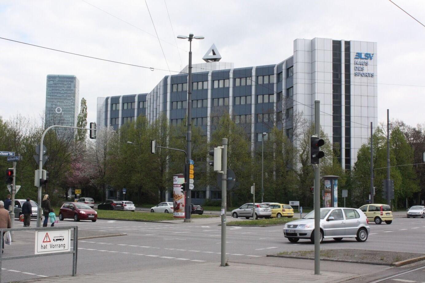 Blick auf ein modernes B&uuml;rogeb&auml;ude mit Glasfassade an einer stark befahrenen Kreuzung mit Verkehrsampeln.
