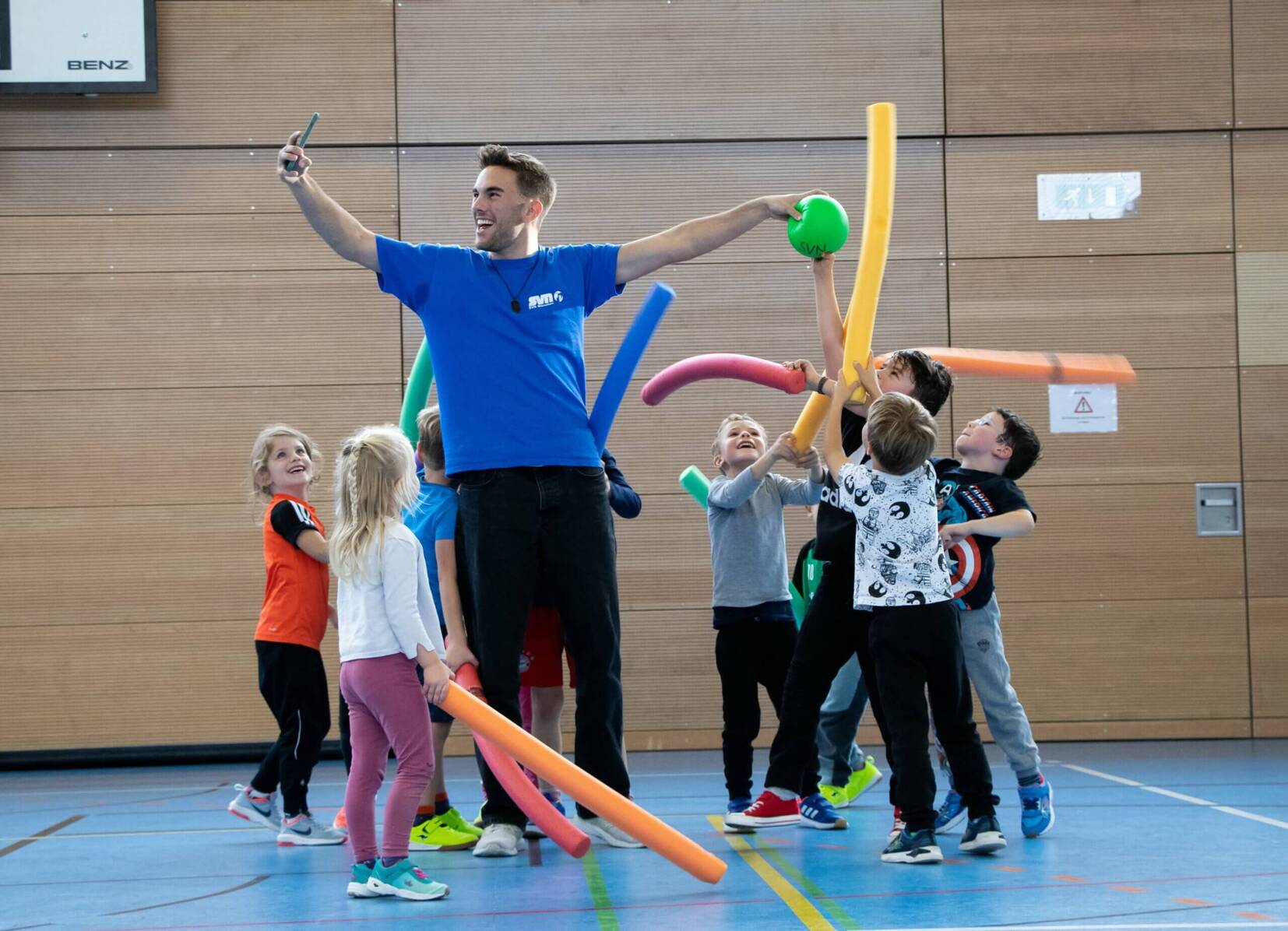 Ein Trainer mit einem Ball steht umgeben von Kindern, die bunte Schwimmnudeln in einer Sporthalle halten.