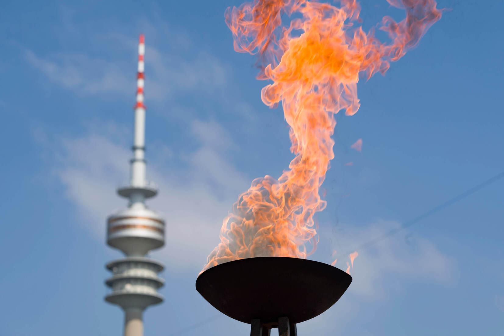 Flamme in einer Schale vor einem Fernsehturm, unter blue Himmel mit wenigen Wolken.