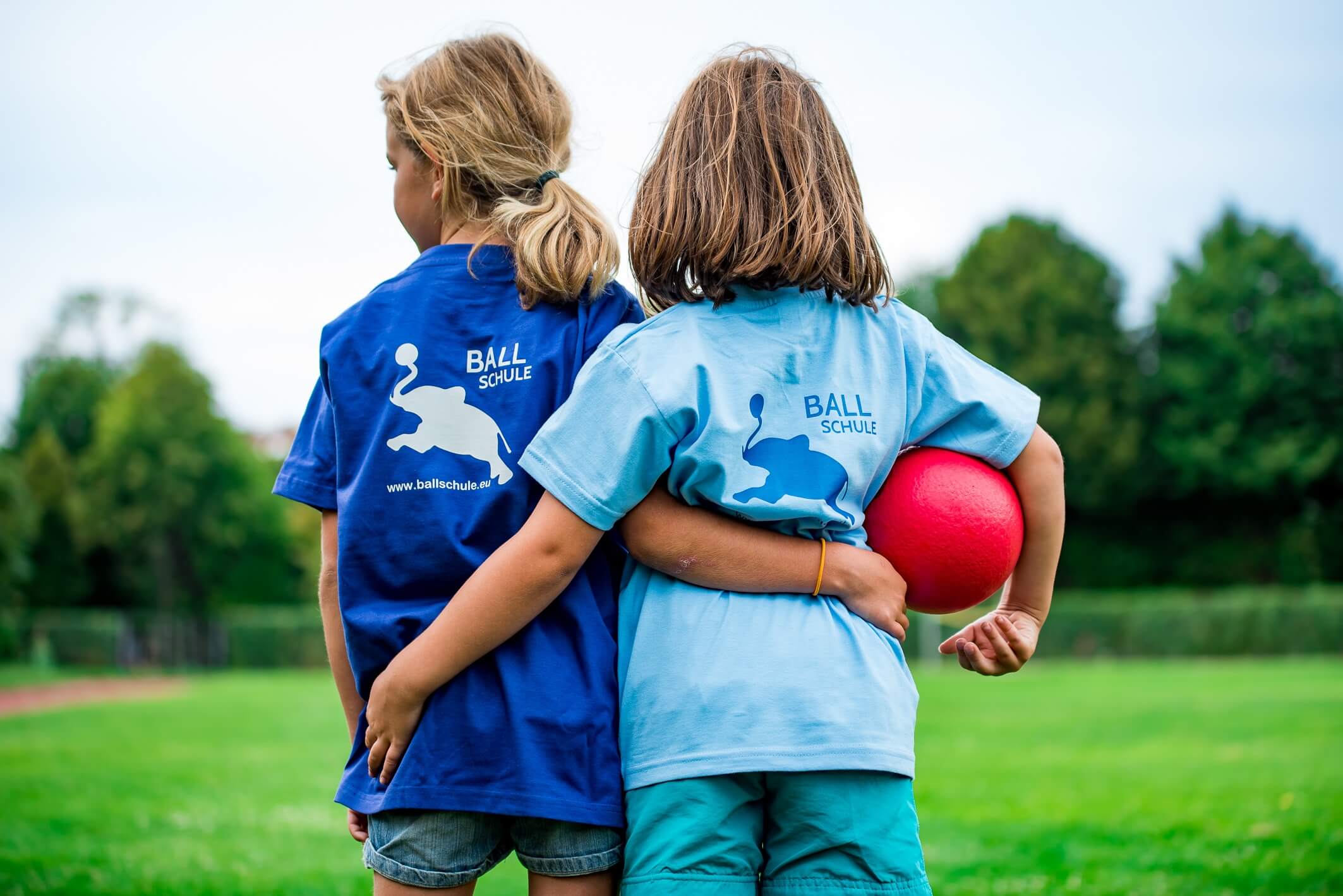 Zwei Kinder stehen R&uuml;cken an R&uuml;cken auf einem gr&uuml;nen Feld, eines h&auml;lt einen roten Ball und tr&auml;gt ein blaues T-Shirt.