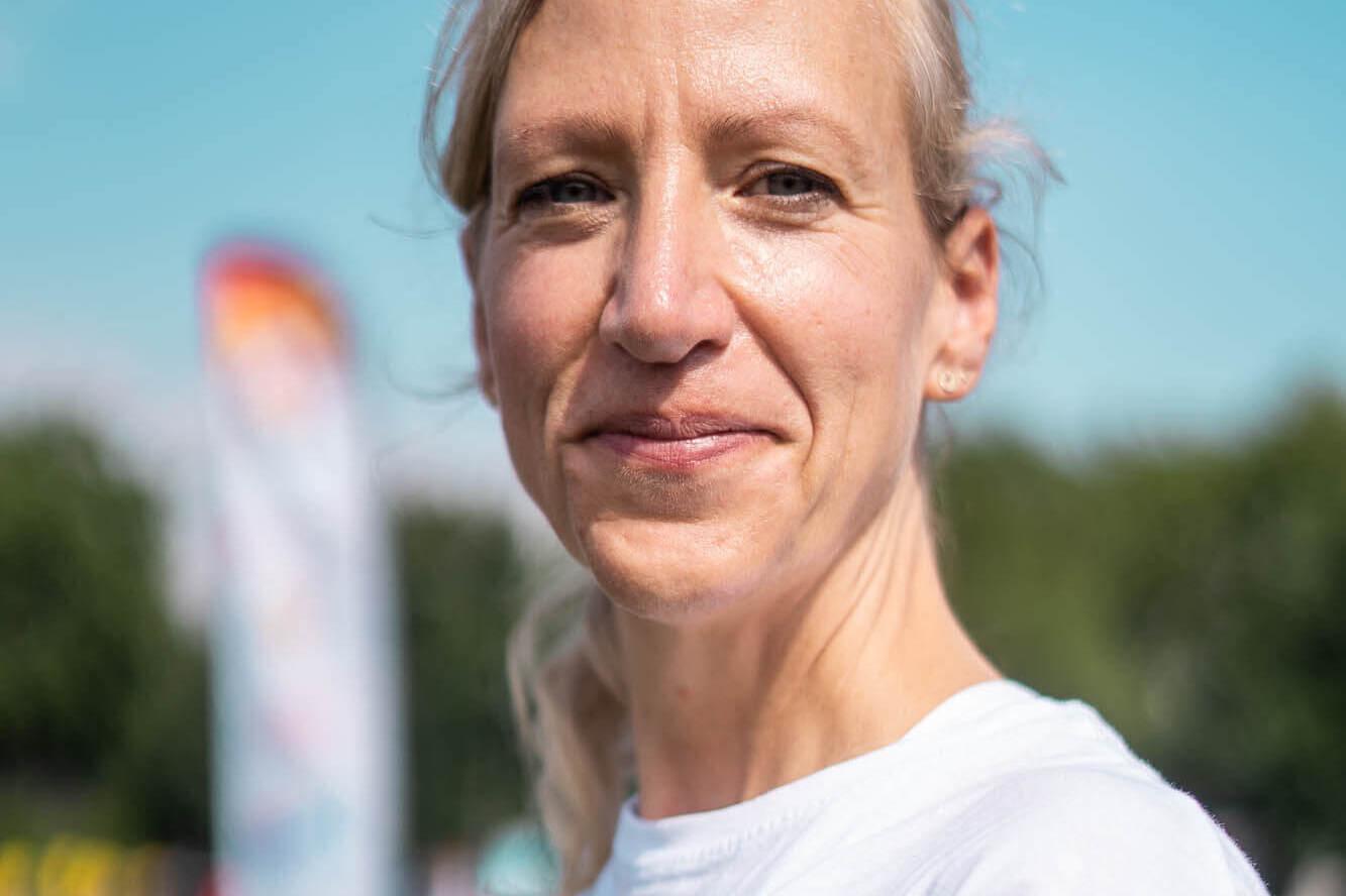 Frau mit blonden Haaren, trägt ein weißes T-Shirt, im Hintergrund eine Sportveranstaltung und in blauer Himmel.
