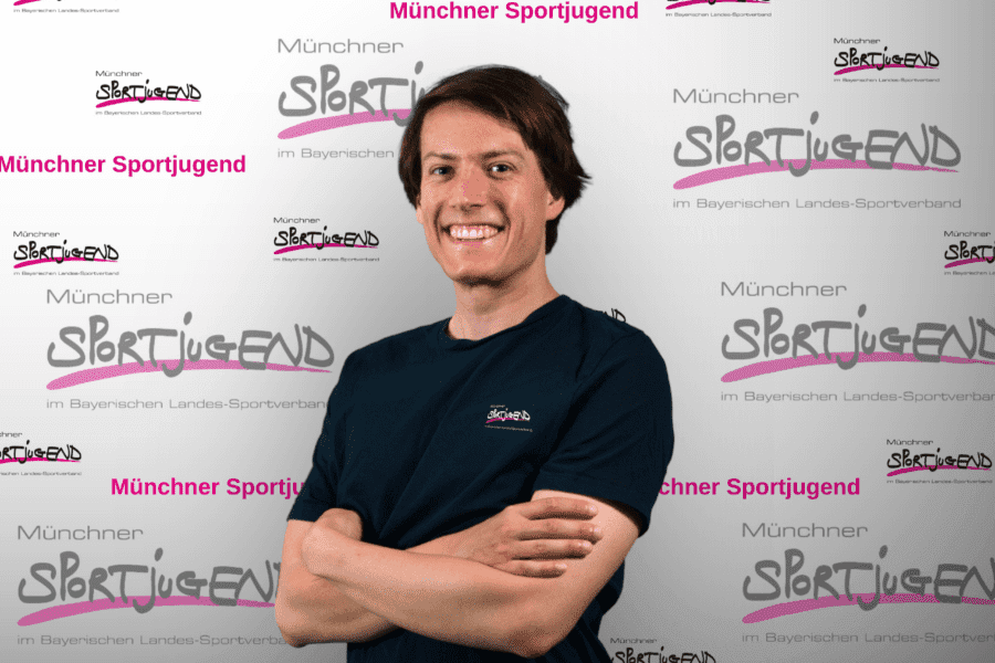 Junger Mann mit lächelndem Gesicht steht vor einem Banner der Münchner Sportjugend. Er trägt ein schwarzes T-Shirt.