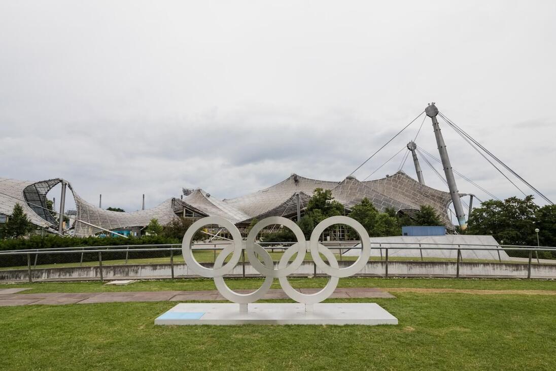 Olympia-Ringe aus weißem Material auf einer Rasenfläche, im Hintergrund das markante Dach der Olympiastätte.