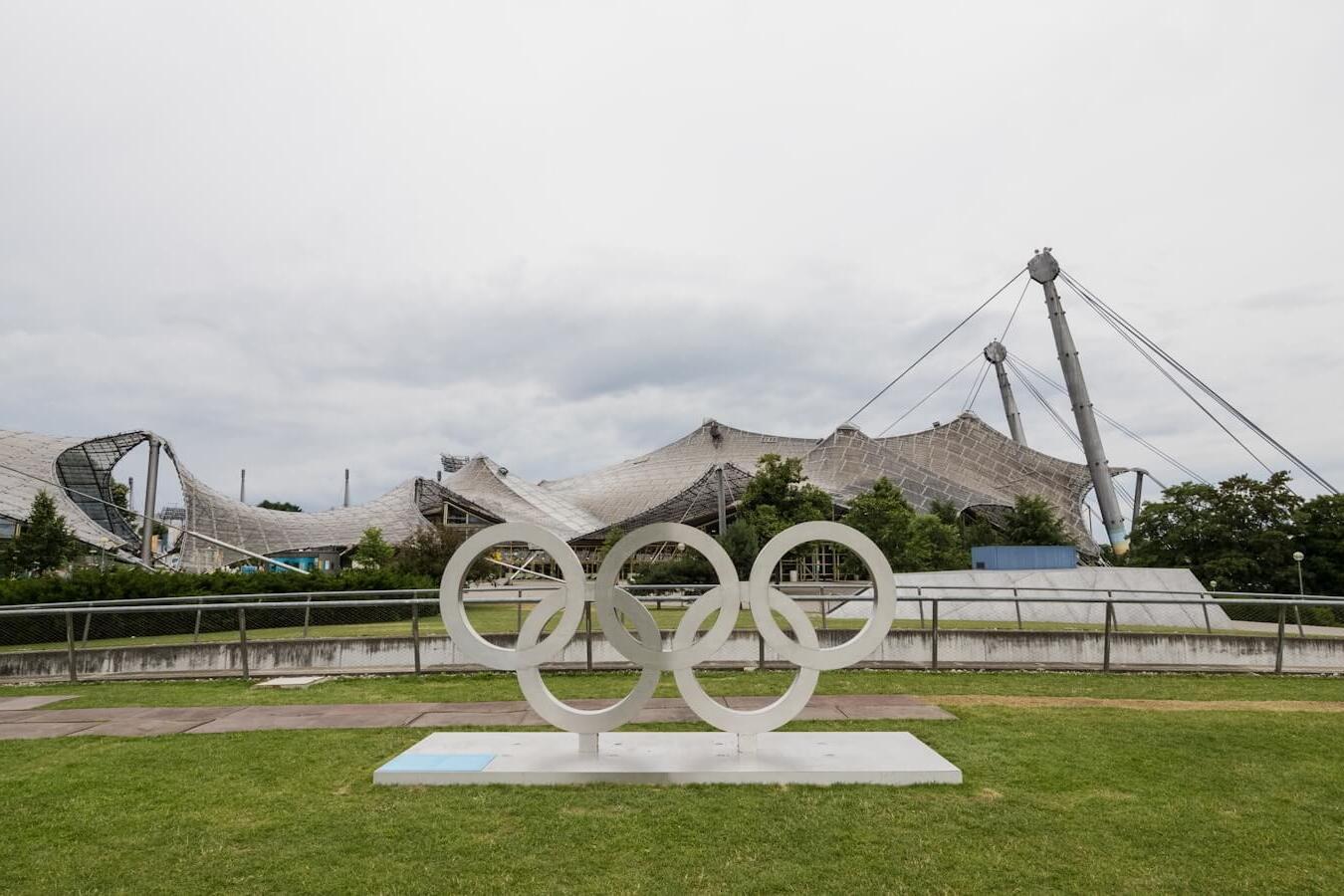 Olympia-Ringe aus weißem Material auf einer Rasenfläche, im Hintergrund das markante Dach der Olympiastätte.