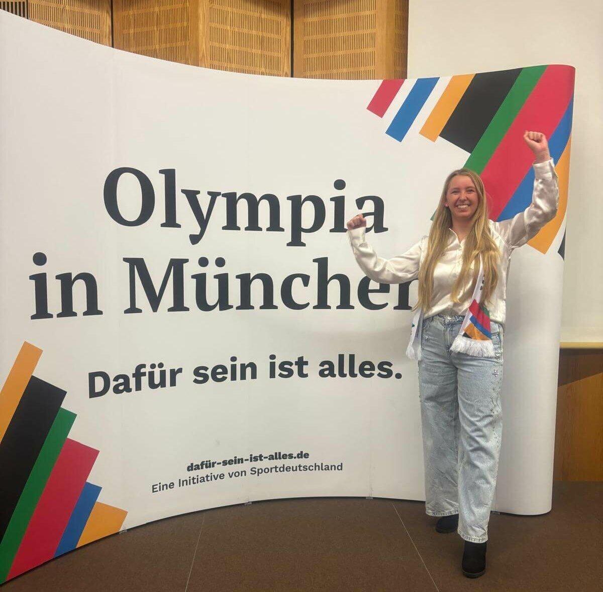 Frau mit langen Haaren steht vor einem Banner mit dem Text &bdquo;Olympia in M&uuml;nchen&ldquo; und hebt siegesbewusst die H&auml;nde.