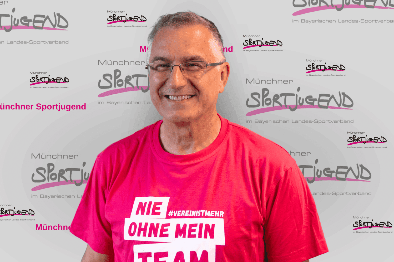 Mann mit Brille und grauen Haaren tr&auml;gt ein pinkes T-Shirt mit dem Schriftzug &bdquo;Nie ohne mein Team&ldquo;.