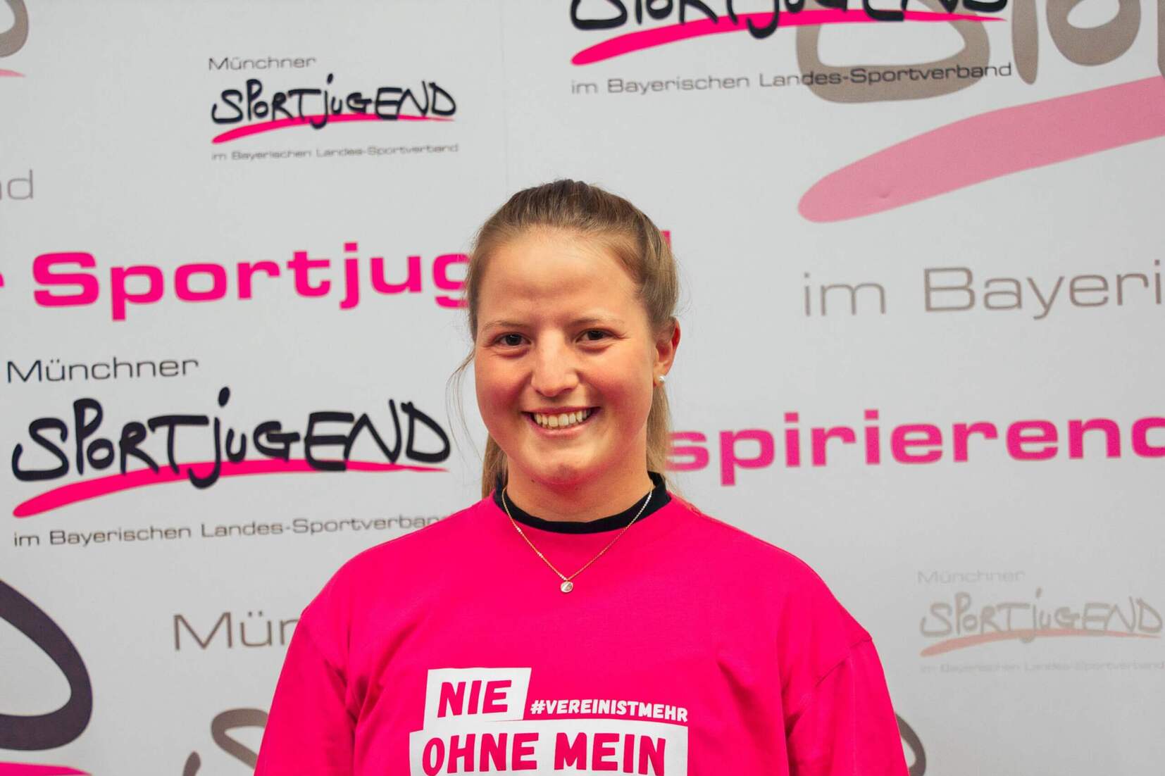 L&auml;chelnde Person in pinkem T-Shirt mit dem Aufdruck &bdquo;Nie ohne mein Team&ldquo; vor einem beschrifteten Hintergrund.