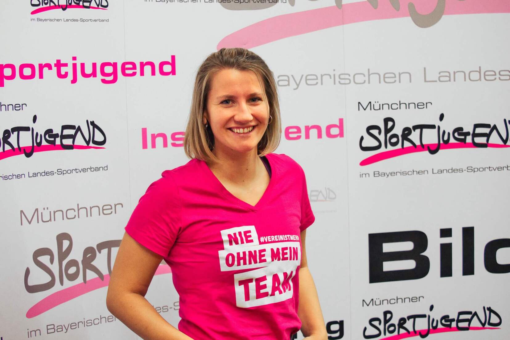 Frau mit kurzem Haar in pinkem Shirt mit Aufschrift &bdquo;NIE OHNE MEIN TEAM&ldquo; vor einem wei&szlig;en Hintergrund mit Logos.