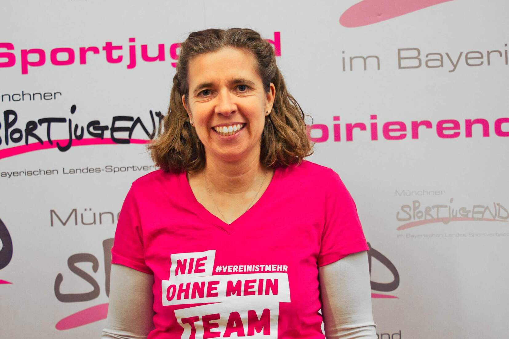 Frau mit lockigem Haar tr&auml;gt ein pinkes T-Shirt mit dem Slogan &bdquo;Nie ohne mein Team&ldquo; vor einem Sportjugend-Hintergrund.