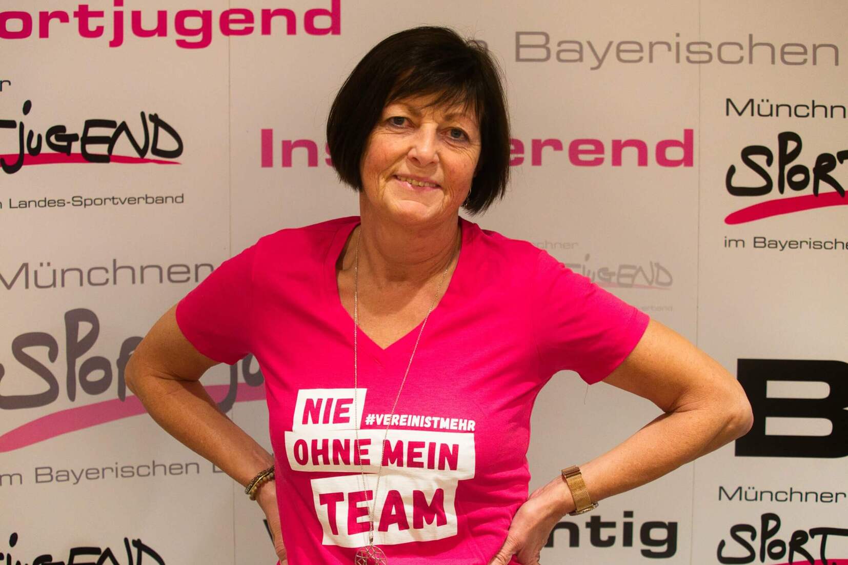 Frau in rotem T-Shirt mit Aufschrift &bdquo;NIE OHNE MEIN TEAM&ldquo; posiert vor einem beschrifteten Hintergrund.