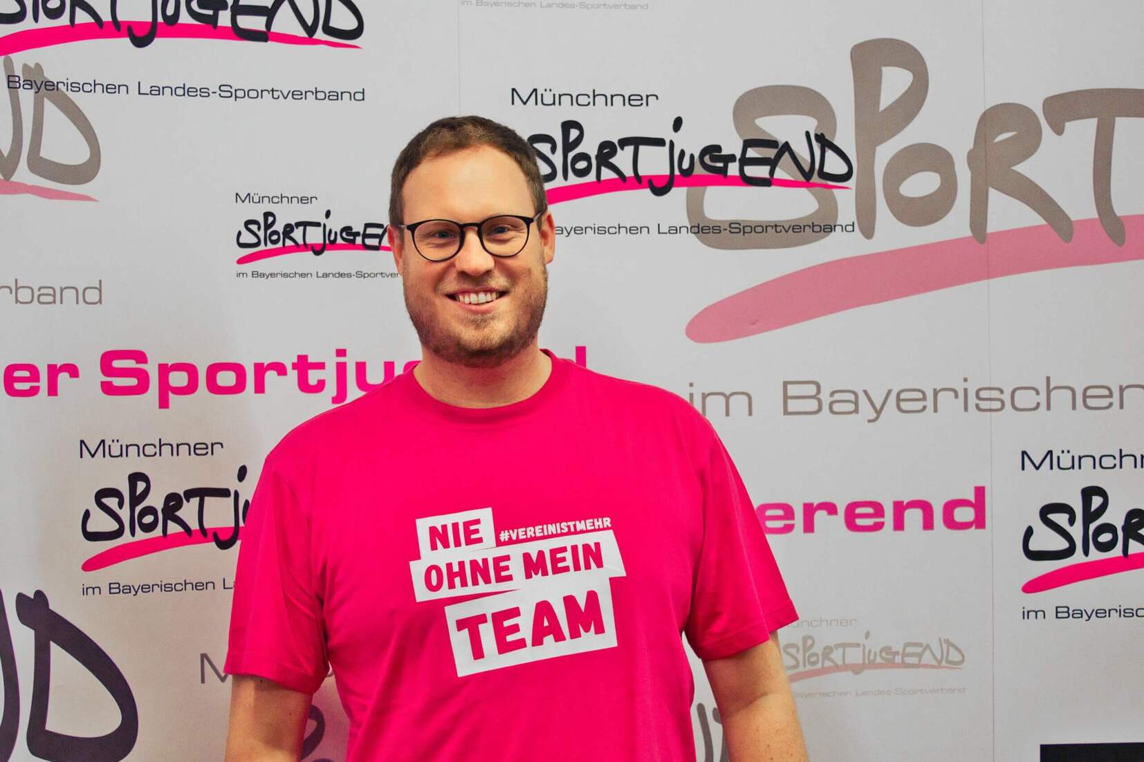 Mann mit Brille in pinkem T-Shirt steht vor einem Banner der M&uuml;nchner Sportjugend.