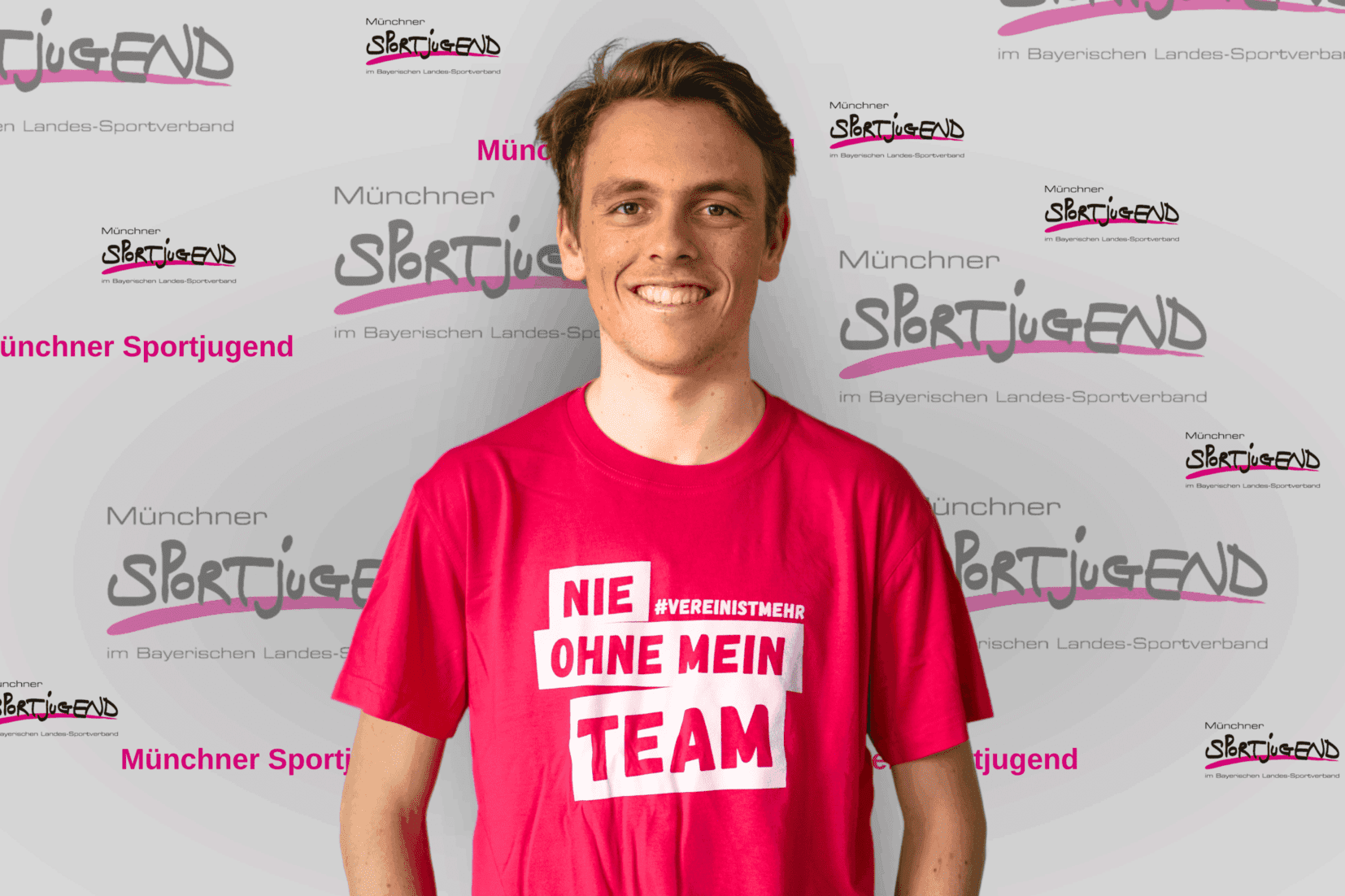 Junger Mann in pinkem T-Shirt mit Aufschrift &bdquo;NIE OHNE MEIN TEAM&ldquo;, vor Hintergrund mit &bdquo;M&uuml;nchner Sportjugend&ldquo;-Logo.