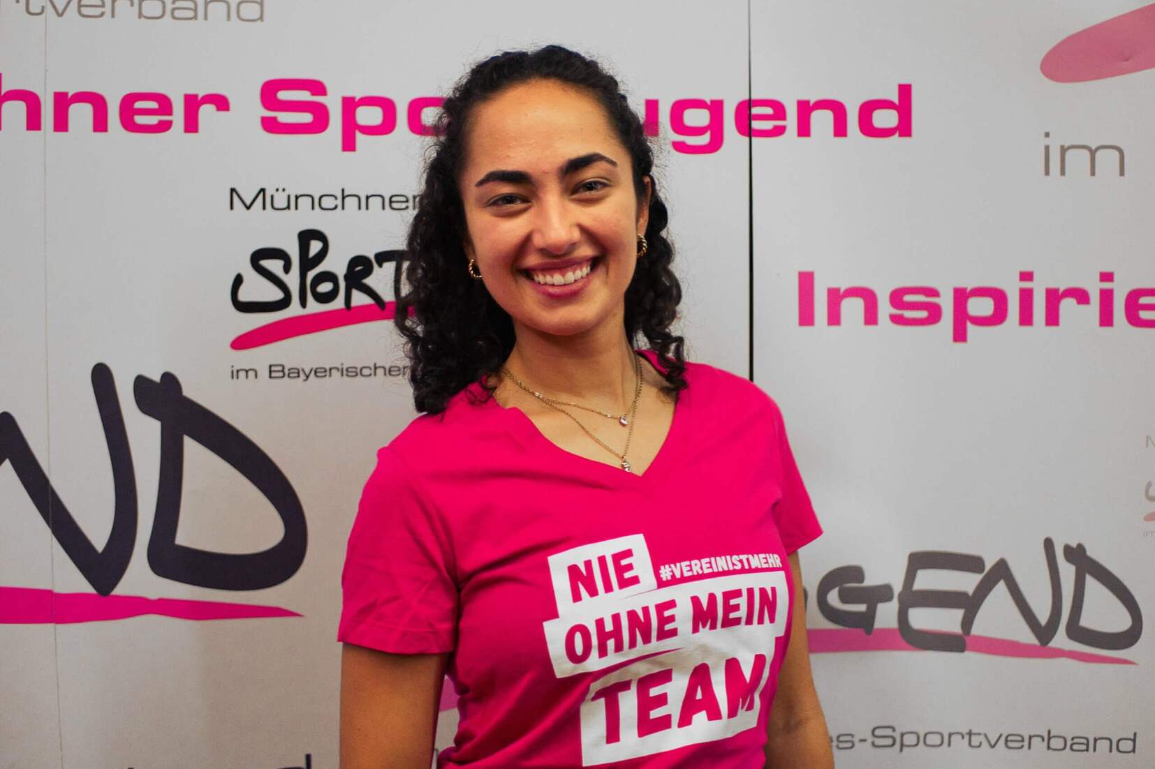 Junge Frau mit lockigem Haar trägt ein pinkes Shirt mit dem Text „Nie ohne mein Team“ vor einem Sportjugend-Hintergrund.