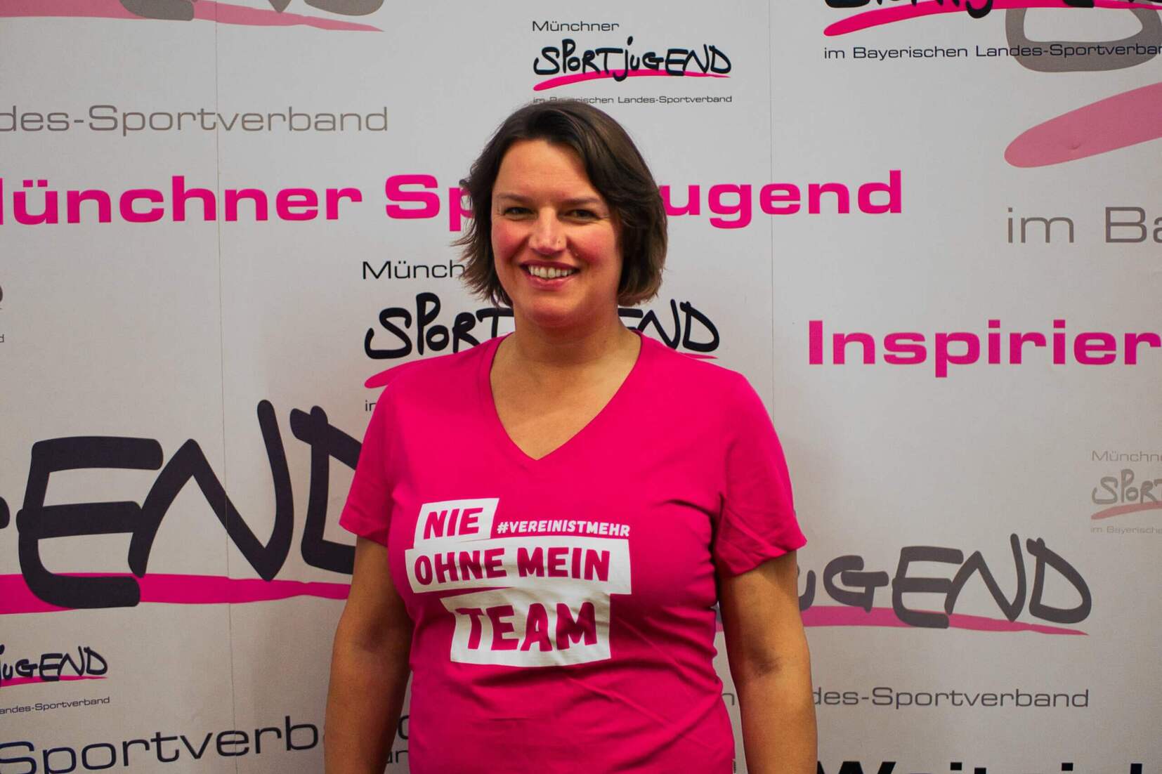 Frau mit kurzem Haar trägt ein pinkes Shirt mit dem Slogan „NIE OHNE MEIN TEAM“ vor einer bunten Wand mit Logos.