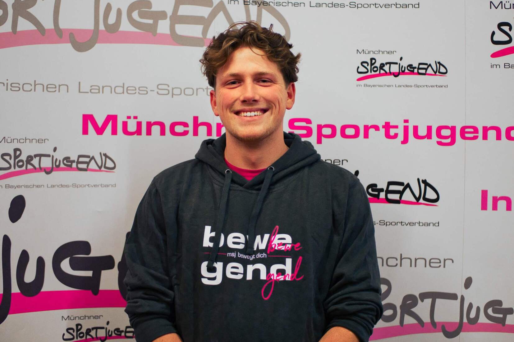 Junger Mann in Sweatshirt lächelt vor einem Banner der Münchner Sportjugend.