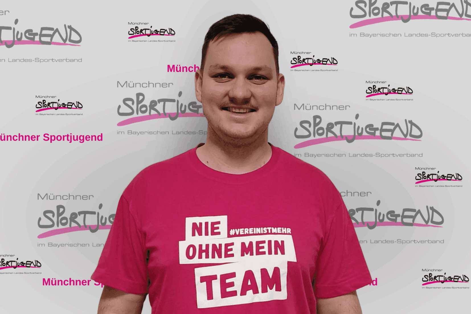 Mann mit kurzem Haar trägt ein pinkes T-Shirt mit Aufschrift "NIE OHNE MEIN TEAM" vor einem Sportjugend-Hintergrund.