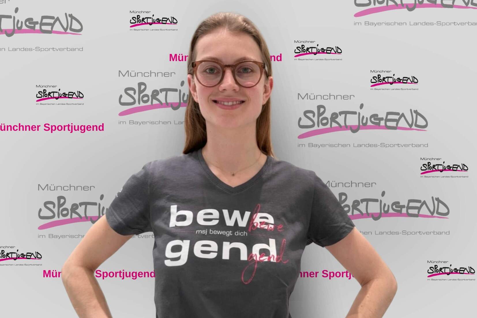 Frau mit Brille steht vor einem Hintergrund mit dem Schriftzug „Münchner Sportjugend“ und trägt ein Shirt mit Aufdruck.