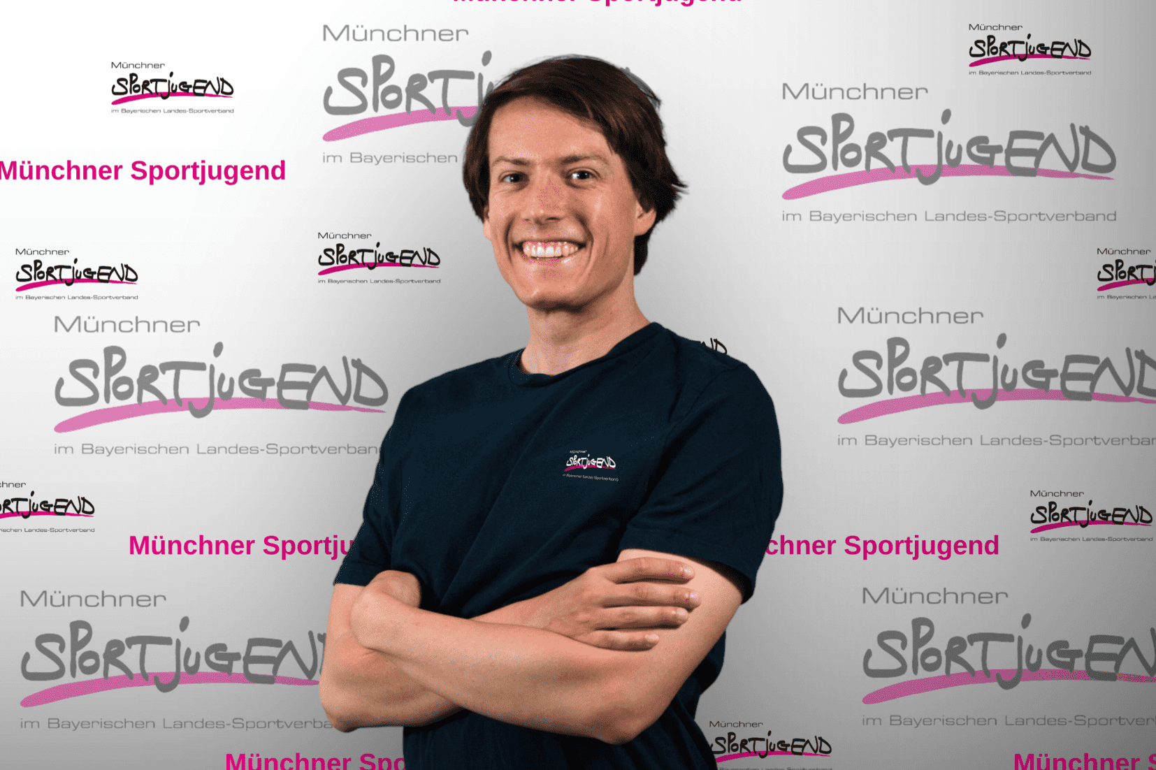Junger Mann mit braunen Haaren und lächelndem Gesicht, steht vor einem Hintergrund mit dem Logo „Münchner Sportjugend“.