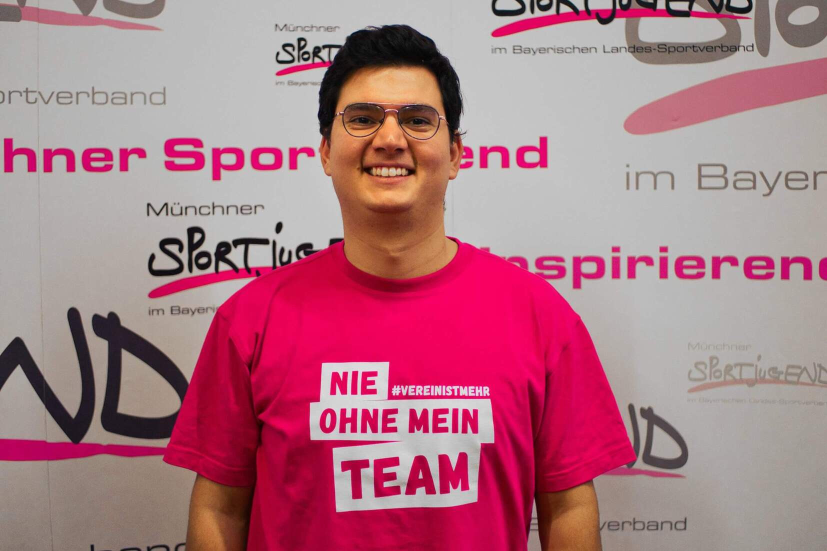 Junger Mann mit Brille tr&auml;gt ein pinkes T-Shirt mit der Aufschrift &bdquo;NIE OHNE MEIN TEAM&ldquo; vor einem bunten Hintergrund.