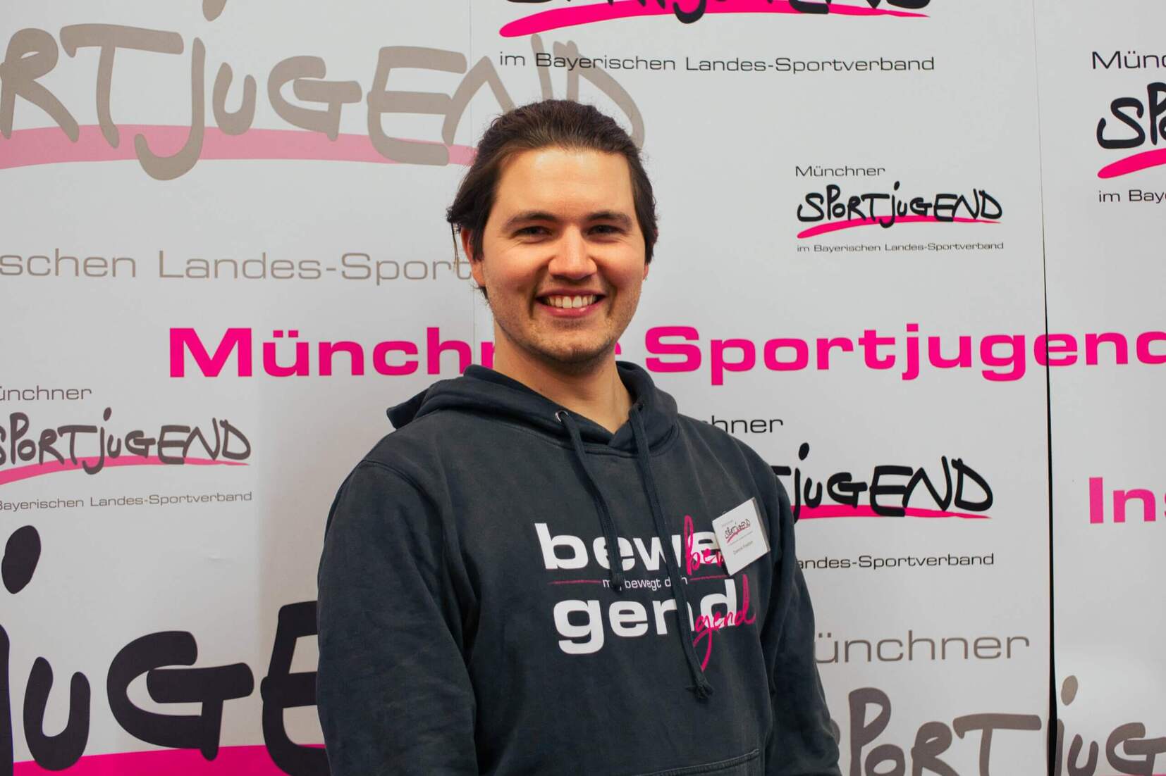 Lächelnder junger Mann mit dunklem Hoodie steht vor einer Wand mit dem Schriftzug "Münchener Sportjugend".