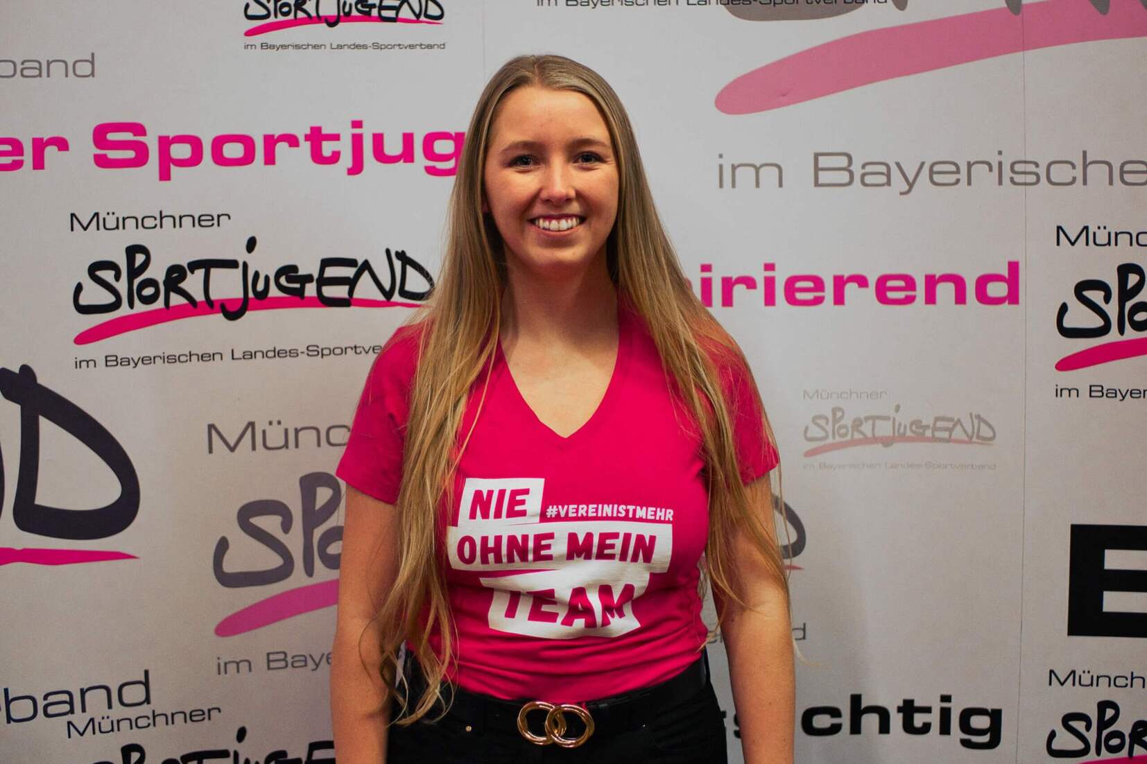 Frau mit langen Haaren in pinkem T-Shirt steht vor einer bunten Wand mit Schriftzügen zu Sport und Teamgeist.