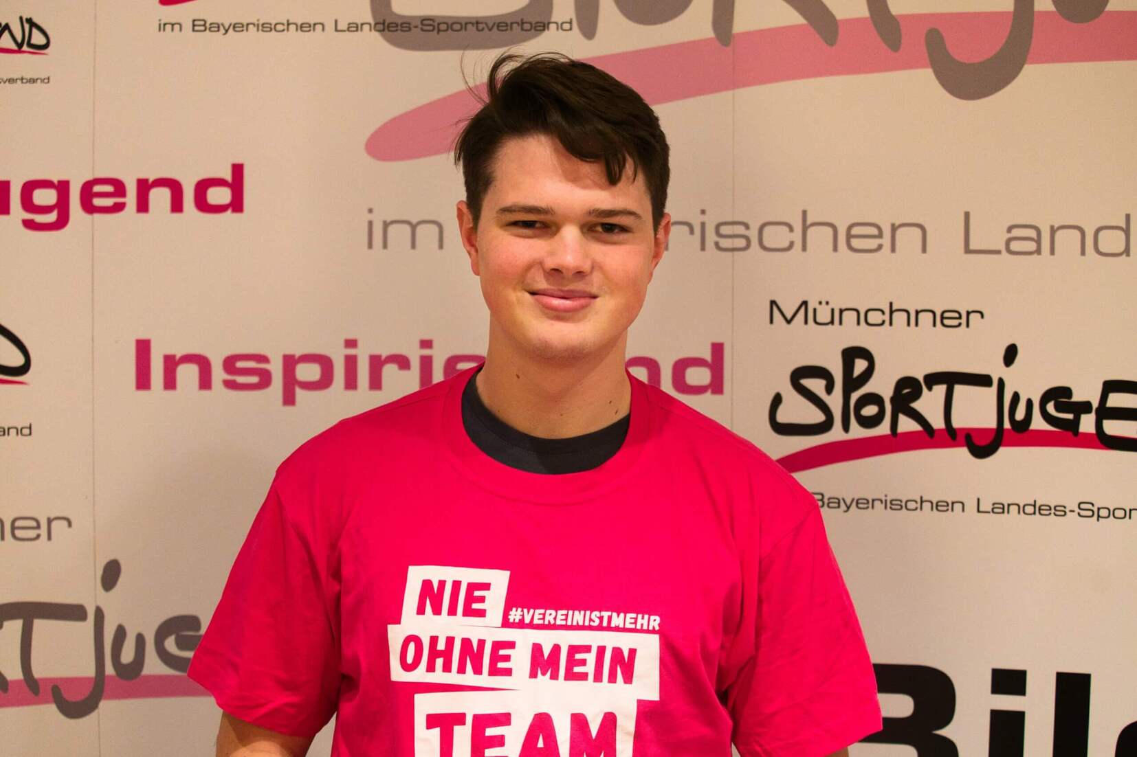 Junger Mann in pinkem T-Shirt mit dem Schriftzug „Nie ohne mein Team“ vor Sportjugend-Hintergrund.