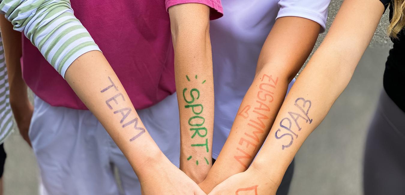 Vier Arme liegen übereinander, mit farbiger Schrift: „TEAM“, „SPORT“, „ZUSAMMEN“, „SPASS“ auf der Haut.
