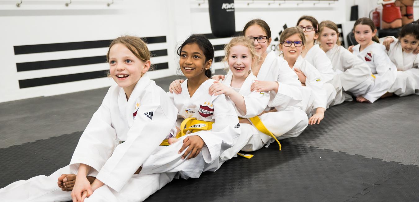 Gruppe von lächelnden Mädchen in weißen Karate-Anzügen sitzt auf einer Matte in einem Trainingsraum.