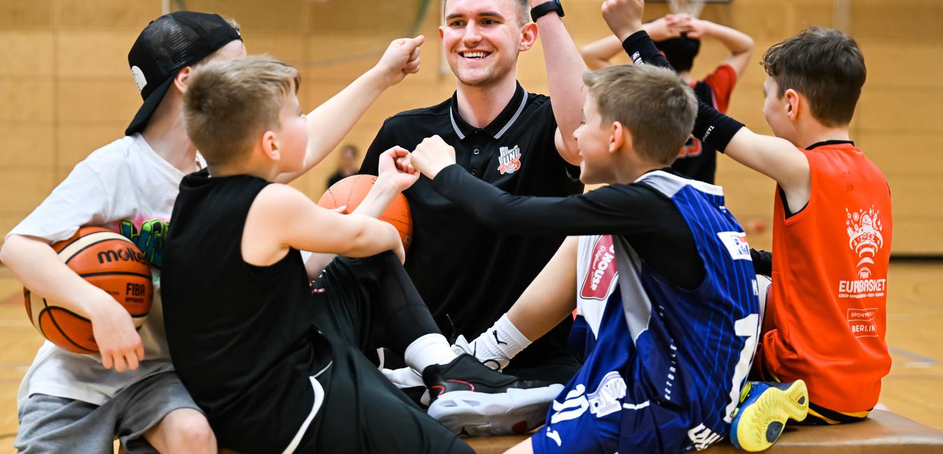 Sechs Kinder und ein Trainer zeigen Freude beim Basketballtraining auf einer Holzbank in einer Sporthalle.