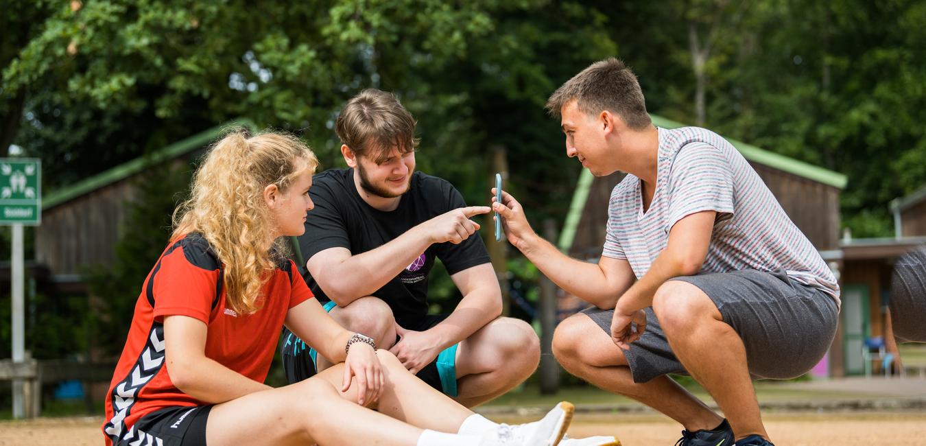 Drei Jugendliche sitzen auf einem Sandplatz und betrachten gemeinsam ein Smartphone. Bäume und Hütten im Hintergrund.