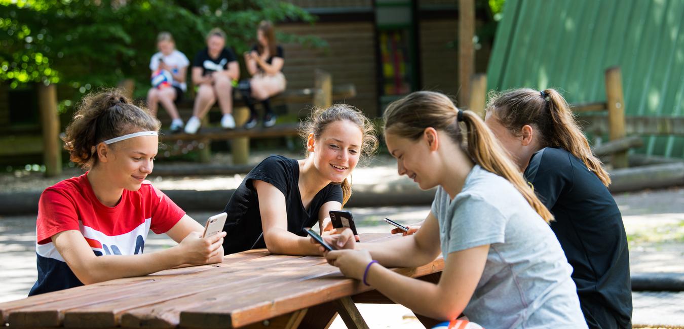 Vier Mädchen sitzen an einem Holztisch und nutzen ihre Smartphones, während im Hintergrund andere Kinder spielen.