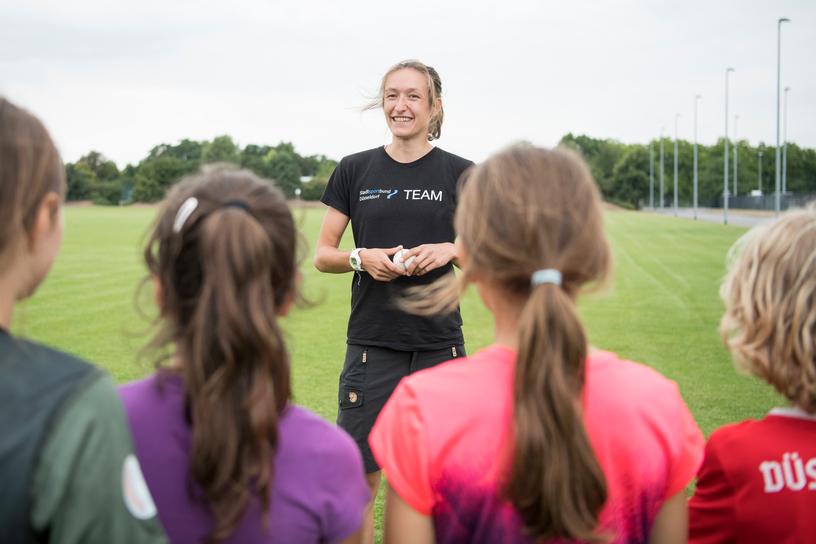 Trainerin spricht mit jungen Sportlerinnen auf einem grünen Fußballfeld unter bewölkten Himmel.