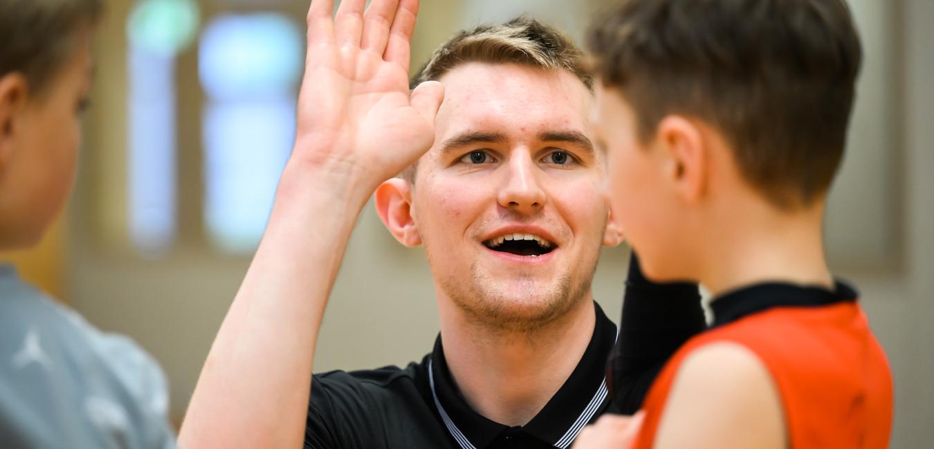 Trainer spricht mit einem jungen Spieler, während er seine Hand hebt, in einer Sporthalle mit Holzwand.